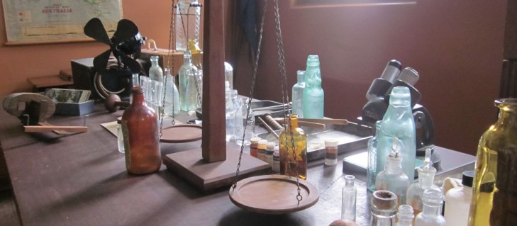 A collection of old bottles, a set of scales, a stereoscope and a bar bell on a wooden desk