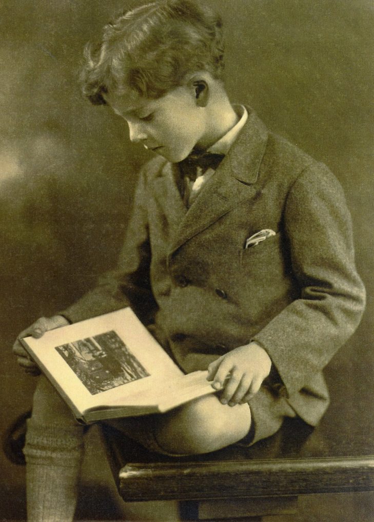 Geoffrey Collins 7yrs dressed in collar and tie ,shorts and long socks sitting on a wooden chair reading a book.