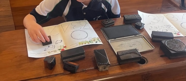 Child pressing a small rectangular rubber stamp onto a page with an image of a clockface and images of numbers