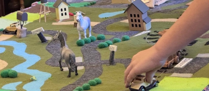 A felt map with 3D toy houses, horses, roads and cars. A child is moving an old fashioned car along a road.